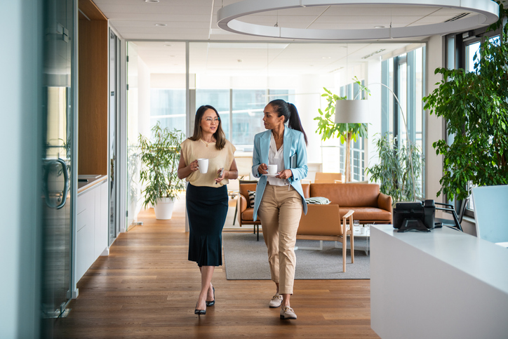 Asian and Hispanic mid-adult businesswomen converse while walking through an office hallway, dressed in formal work attire and carrying documents.